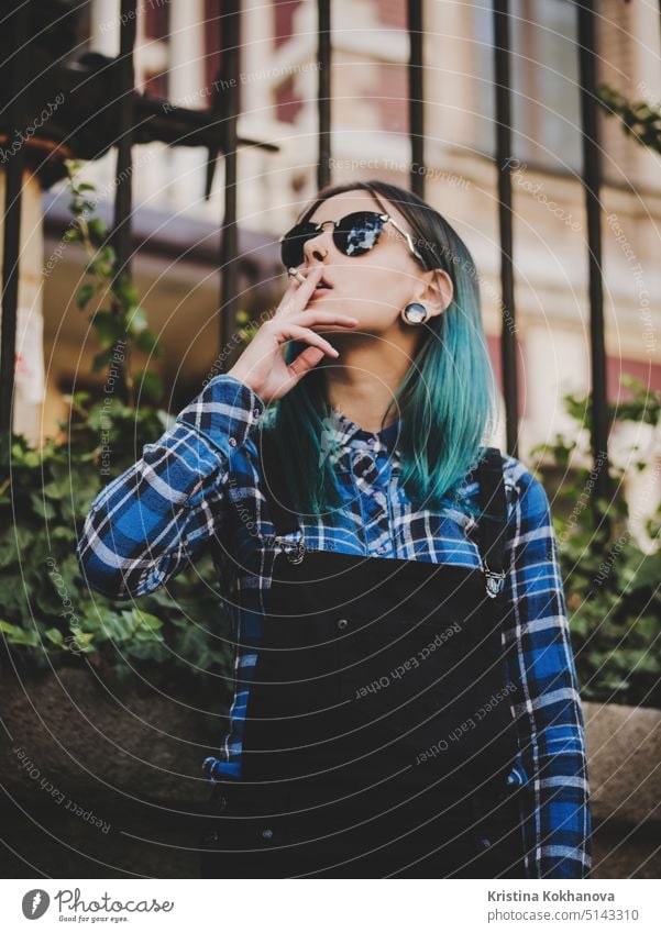 Young attractive punk girl smoking cigarette. Hipster with blue unusial dyed hair, piercing in nose, violet lenses, ears tunnels on empty European old street.