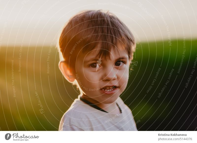 Portrait of smiling kid standing in fresh green field. Lovely toddler boy explores plants, nature in spring. Childhood, future, agriculture, ecology concept