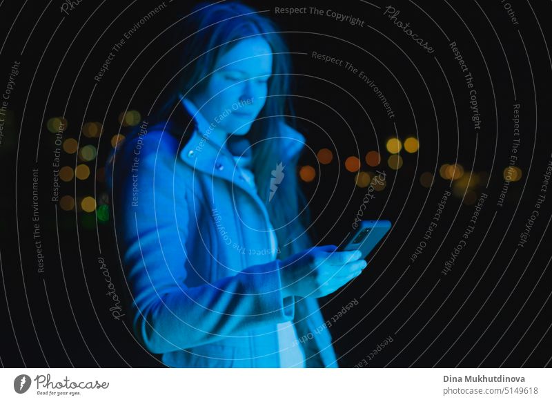 woman holding mobile phone, texting and using social media in blue neon light in city at night. Real people using technology. Woman standing outside with smartphone in her hands.