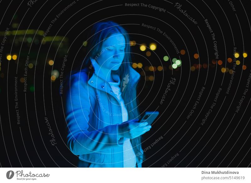 woman holding mobile phone, texting and using social media in blue neon light in city at night. Real people using technology. Woman standing outside with smartphone in her hands.