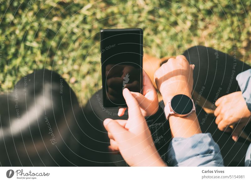 happy friends sharing multimedia content online with their smartphones and smartwatch, two beautiful Latina women sitting outdoors advertising atmospheric bokeh