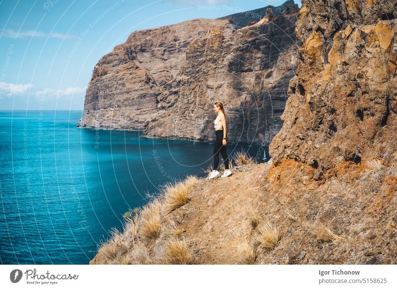 Independent relaxed calm slim girl tourist stands on the edge of a cliff with amazing ocean and sky view. Los Gigantes mountain ranges. Tenerife. Santiago del Teide. Canary Islands, Spain.