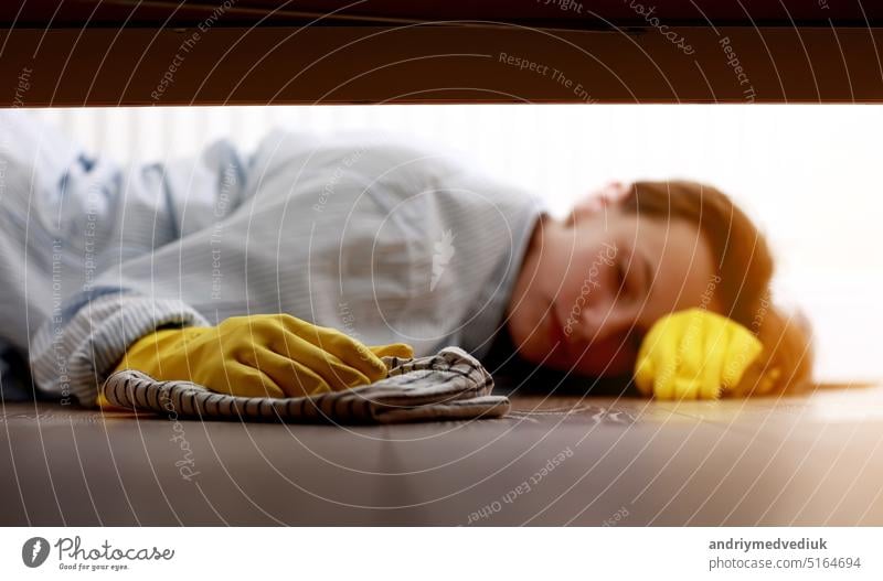 Close up of young beautiful woman with yellow rubber gloves and rag is cleaning floor under bed at home. Concept of cleaning and disinfection of surfaces, housekeeping, lifestyle, routine