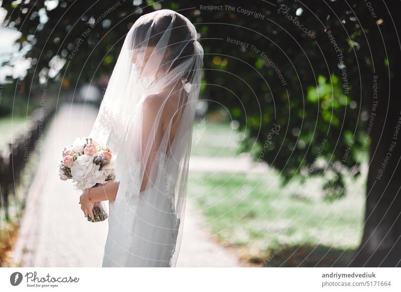 Beautiful bride in bridal gown with bouquet and lace veil on the nature. Beautiful model girl in a white wedding dress. Female portrait in the park. Woman with hairstyle. Cute lady outdoors