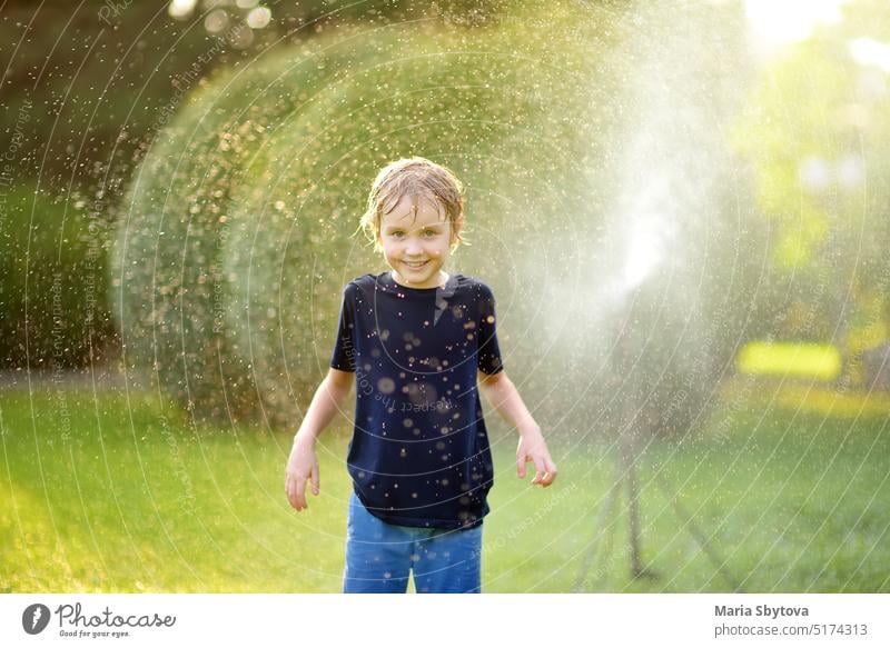 Funny little boy playing with garden sprinkler in sunny backyard. Elementary school child laughing, jumping and having fun with spray of water. Summer outdoors activity for kids.