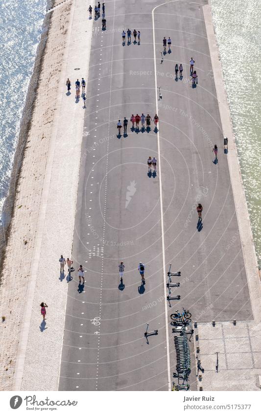 aerial image of groups of people in the distance strolling along a pedestrian street surrounded by canals with water on a summer day, some scooters parked on the street