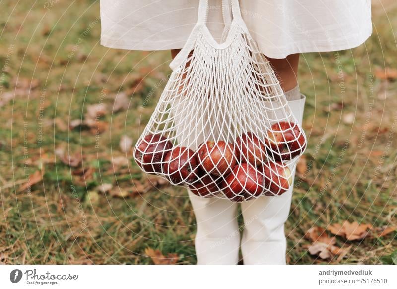 String bag with red apples. View of female legs in white boots walking through the autumn garden with eco bag in hands. Zero waste concept. No plastic life. Healthy fitness lifestyle. Recycling waste