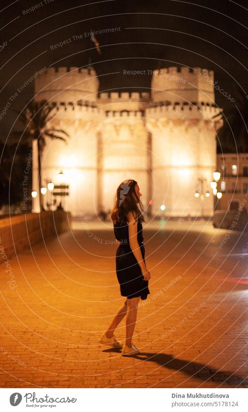 Happy woman walking at night in European town in front of a illuminated castle, smiling. Happy woman walking in the historic center of the city with lights. Smiling happily, going to the party. Young and beautiful female tourist in Spain.