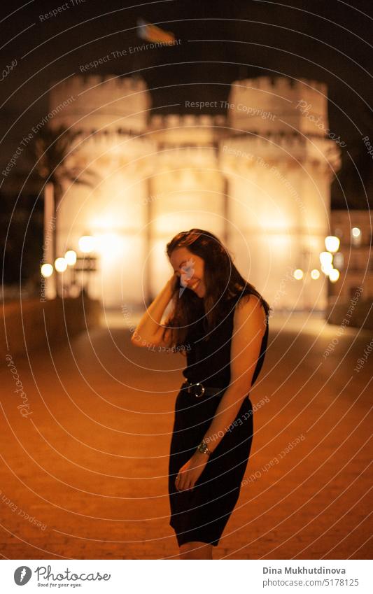 Happy woman walking at night in European town in front of a illuminated castle, smiling. Happy woman walking in the historic center of the city with lights. Smiling happily, going to the party. Young and beautiful female tourist in Spain.