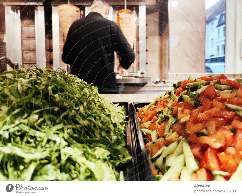 Green salad and tomato cucumber salad in front of two kebab skewers in a snack bar with an employee standing at the kebab skewer Snack bar Eating Meat Veal