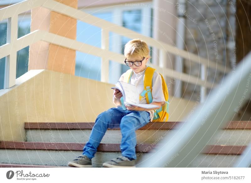 Smart little child sitting and reading on the stairs of school building. Quality education for children. Portrait of funny nerd schoolboy with big glasses. Vision problems. Back to school concept.