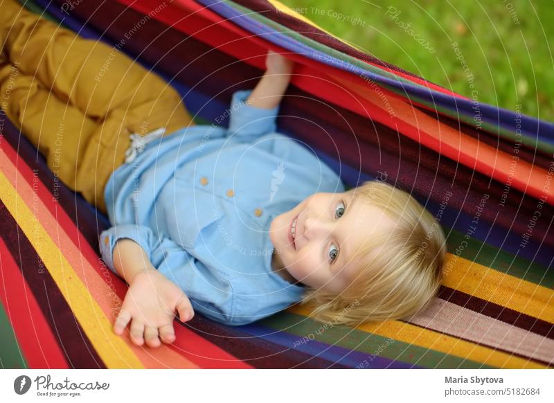 Cute little blond white boy enjoy and having fun with multicolored hammock in backyard or outdoor playground. Summer outdoors active leisure for kids. Child relaxing and swinging in hammock.