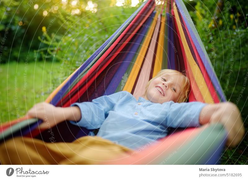 Cute little blond white boy enjoy and having fun with multicolored hammock in backyard or outdoor playground. Summer outdoors active leisure for kids. Child relaxing and swinging in hammock.