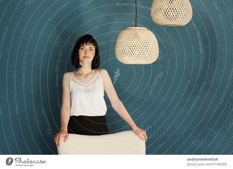 Millennial young brunette woman looking at camera and standing in creative office with grange blue green wall, soft chair and rattan lamps . Successful self-confident female designer. copy space