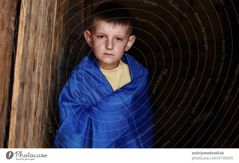 sad Ukrainian small child stands outdoors supporting homeland, little kid covered in national flag. Protesting against Russian war in Ukraine, armed conflict, homeless, crisis, portrait