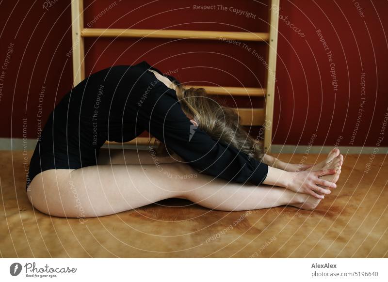 a young blonde slim woman in black bodysuit is doing gymnastics on a gym mat in front of a red wall with wall bars Woman youthful Slim Athletic pretty Smiling