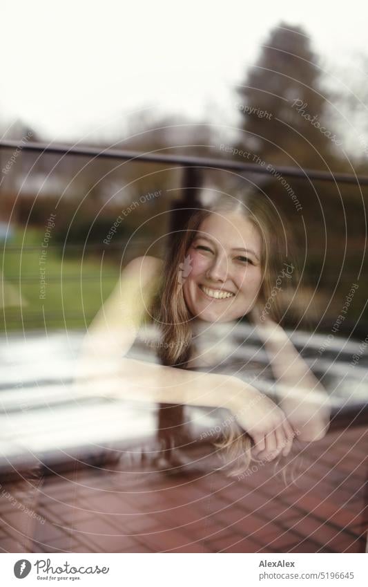 a young blonde slim woman in black dress is lying on the couch behind the balcony window pane and smiling into the camera Woman youthful Slim Athletic pretty