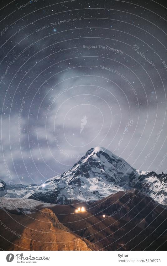 Stepantsminda, Georgia. Night Starry Sky With Glowing Stars Above Peak Of Mount Kazbek Covered With Snow. Famous Gergeti Church In Night Lightning. Beautiful Georgian Landscape In Late Autumn