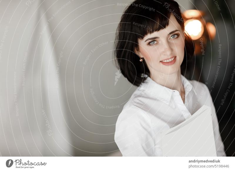 beautiful young woman with dark short hair holding a magazine, book with mock up. girl in a white shirt and black leather skirt standing in studio reading