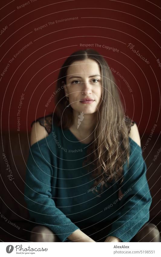 Portrait of young brunette woman with off shoulder sweater sitting in front of red wall Face Face of a woman Woman Young woman pretty daintily Brunette Bright