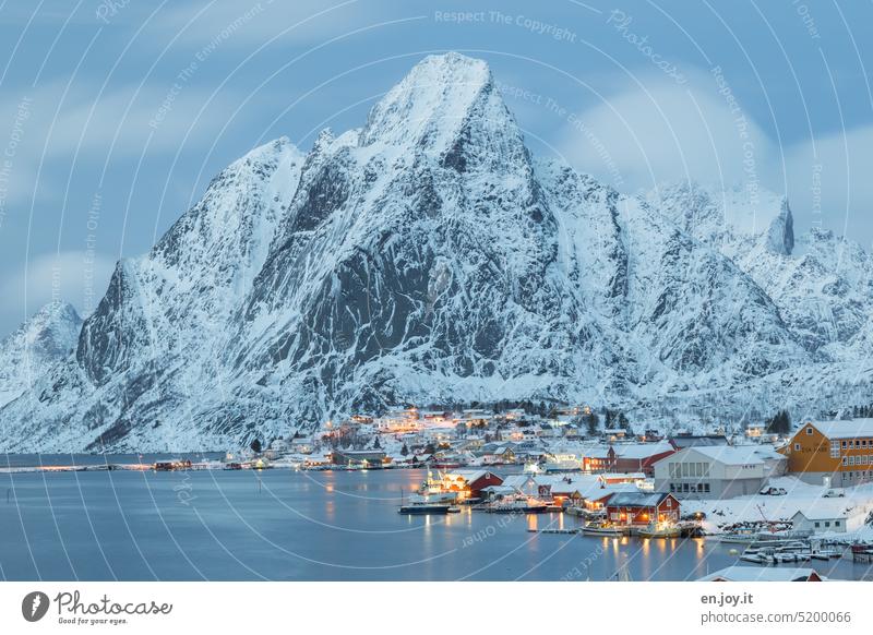 Fishing village on fjord in front of mountain in snow Reine Reinefjorden Norway Lofotes Mountain Snow Winter Village Hut White Sky blue hour Evening Twilight