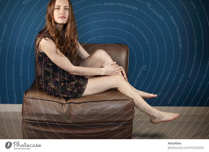 Portrait of a young brunette woman in front of petrol / blue green wall barefoot and slanting sitting on a brown leather armchair Face Face of a woman Woman