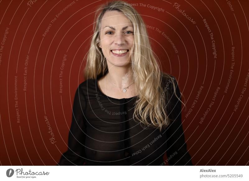 Portrait of blonde long haired woman in black sheer blouse in front of red wall Woman portrait Blonde Long-haired Smiling Looking into the camera Slim daintily