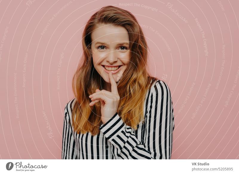 Pretty smiling young girl looking at camera touching lips while standing against pink wall woman positive happy ginger demonstrating female pretty cute redhead