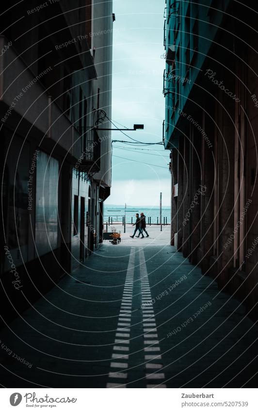 View through a street canyon on two passers-by, behind them the Mediterranean Sea Urban canyon Tunnel Passers-by Pedestrian somber Tall Ocean vacation actuality