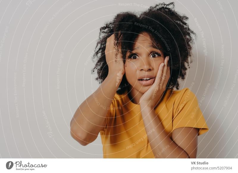 Shocked and nervous dark skinned female student holding hands on head and gasping with worried face frustrated unhappy bright tension not sad studio copyspace