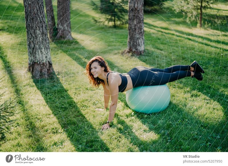 Outdoor shot of active brunette woman exercises with fitness ball poses at green grass, dressed in active wear, enjoys sunny day and fresh air in park, keeps fit, makes gymnastics exercises.