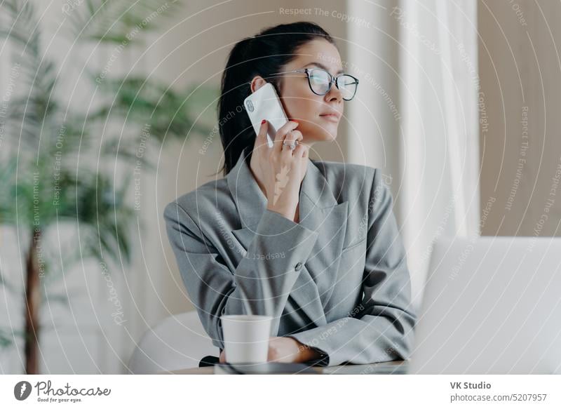 Photo of woman copywriter has telephone talk, looks aside, chats about ideas for new publication, dressed formally, poses in front of laptop computer in coworking space, discuss working process