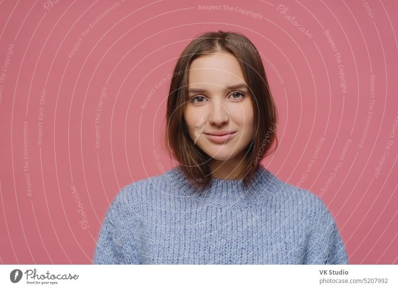 Beautiful adult woman with dark hair, has calm face expression, wears knitted sweater, poses at studio against pink background, thinks about something pleasant during winter time, has tender look