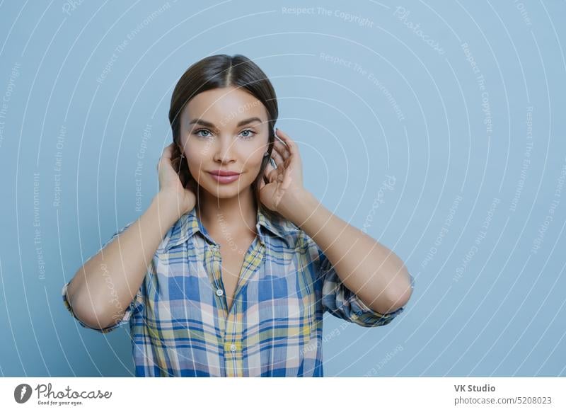 Portrait of beautiful dark haired European woman has healthy glowing skin, make up, dressed in casual checkered shirt, looks directly at camera, stands against blue background, being in good mood