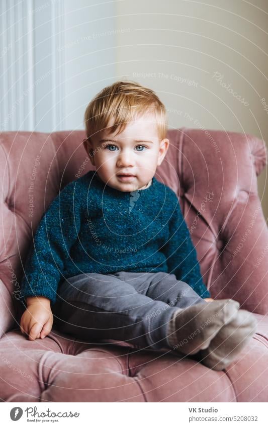 Vertical portrait of innocent little child with blue eyeys and plump cheeks, looks directly into camera, wait for children as has birthday, sits in comfortable pink armchair in living room. Children