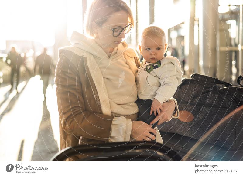 Motherat travelling with his infant baby boy child, walking, pushing baby stroller and luggage cart in front of airport terminal station. vacation trip family