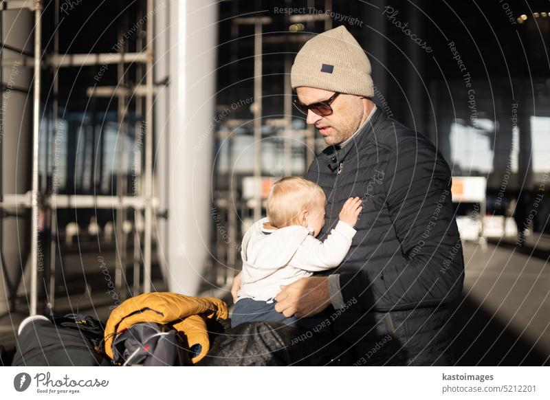 Fatherat comforting his crying infant baby boy child tired sitting on top of luggage cart in front of airport terminal station while traveling wih family.