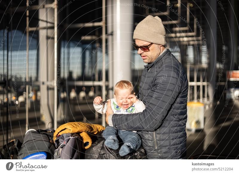 Fatherat comforting his crying infant baby boy child tired sitting on top of luggage cart in front of airport terminal station while traveling wih family.