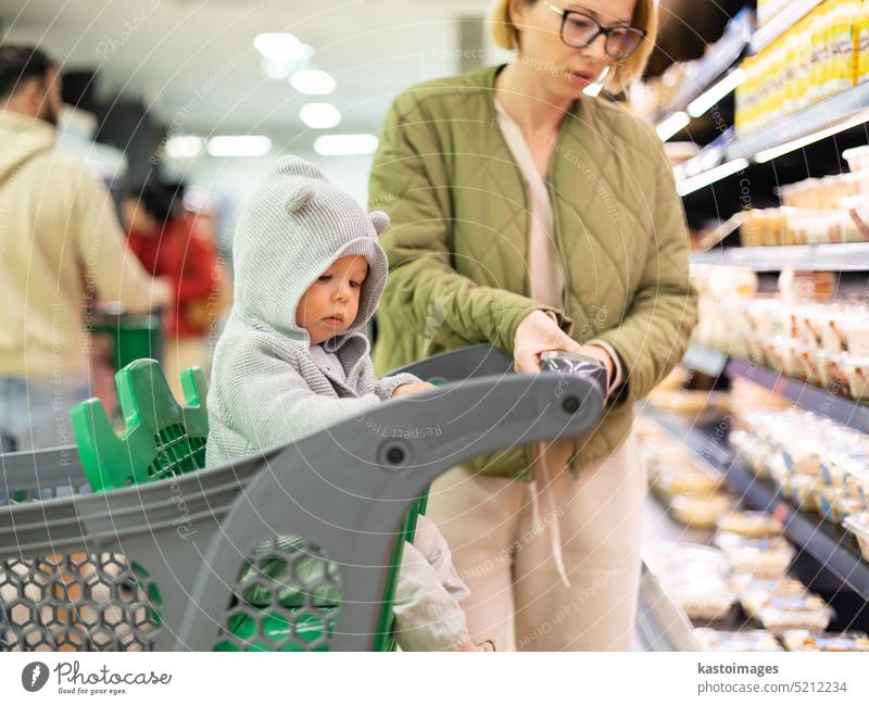 Casualy dressed mother choosing products in department of supermarket grocery store with her infant baby boy child in shopping cart. woman young customer retail