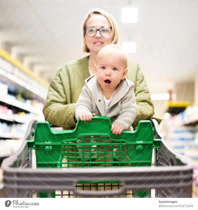 Mother pushing shopping cart with her infant baby boy child down department aisle in supermarket grocery store. Shopping with kids concept. woman mom young