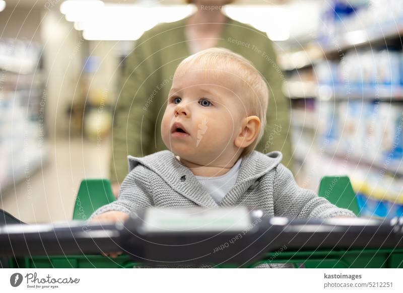 Mother pushing shopping cart with her infant baby boy child down department aisle in supermarket grocery store. Shopping with kids concept. woman mom young