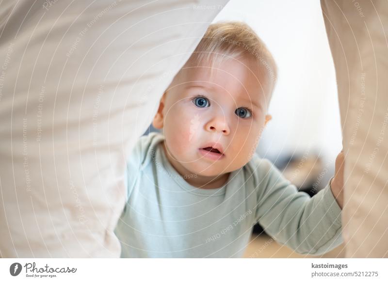 Portrait of adorable curious infant baby boy child taking first steps holding to father's pants at home. Cute baby boy learning to walk. portrait standing small