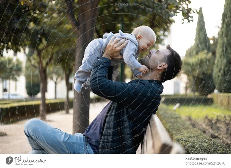 Father holding and lifting his cute infant baby boy child sitting on wooden bench in urban city park. Dad and son enjoing a pristine moment of happiness, smiling and laughing.