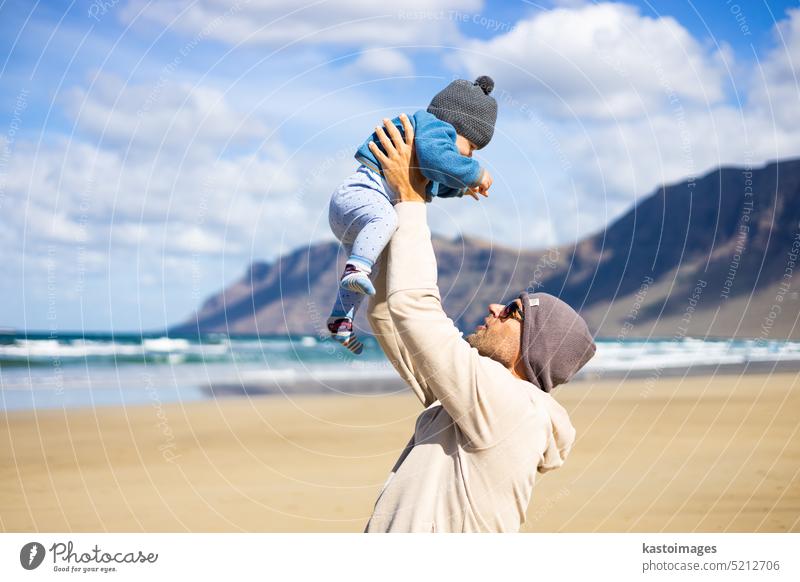 Father enjoying pure nature holding and playing with his infant baby boy son in on windy sandy beach of Famara, Lanzarote island, Spain. Family travel and parenting concept.