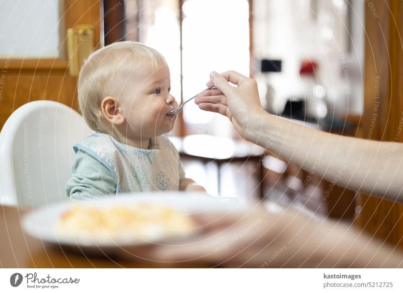 Mother spoon feeding her infant baby boy child sitting in high chair at the dining table in kitchen at home food mother care family hungry adorable beautiful