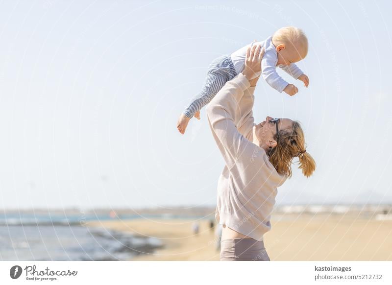 Mother enjoying summer vacations holding, playing and lifting his infant baby boy son high in the air on sandy beach on Lanzarote island, Spain. Family travel and vacations concept.