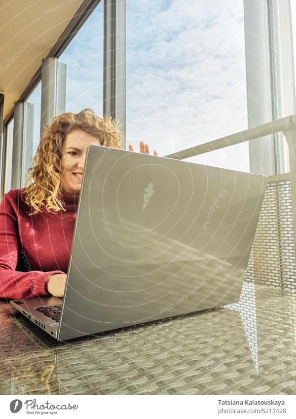 A blonde with curly hair communicates via video link on a laptop sitting in front of a window woman Home office Remote work Working from home Job Mobile working