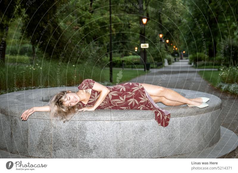 A gorgeous blonde girl is enjoying time in a botanical garden. Wearing a fine summer dress. Showing her beautiful long legs and a big smile. A relaxing time with beauty in a park.