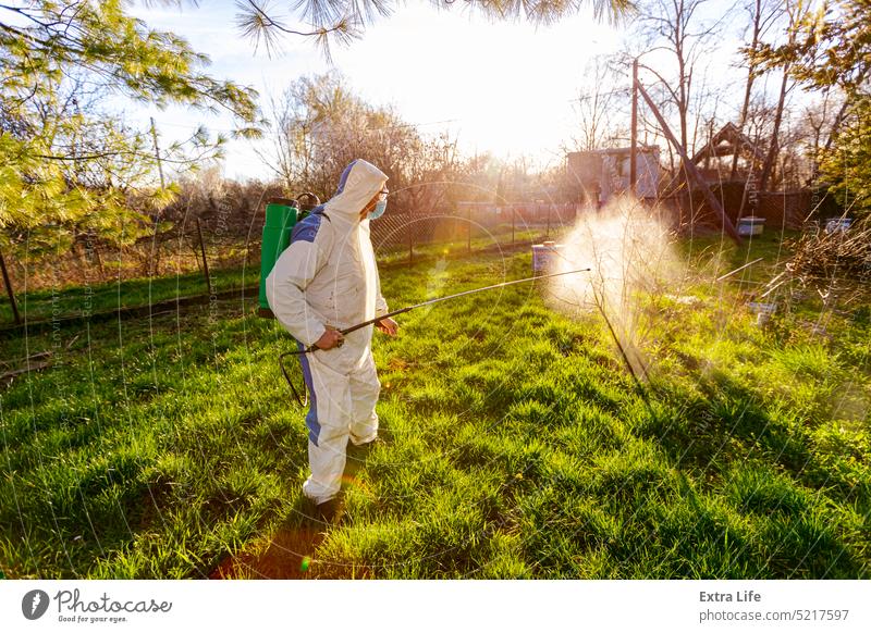 Gardener in protective overall sprinkles fruit trees with long sprayer in orchard Aerosol Agricultural Agriculture Backlight Biochemical Biohazard Blowout