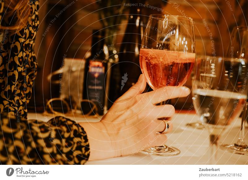 Hand holding glass with red drink, in front of it wine glass, at table in restaurant in evening lighting Glass Beverage splash Ring Wine glass Restaurant